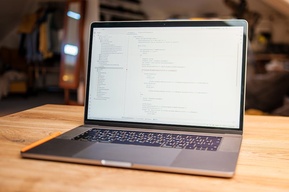 A close-up view of a laptop placed on a wooden table in an indoor setting. The laptop screen displays lines of code with syntax highlighting, indicating programming or software development work. The k