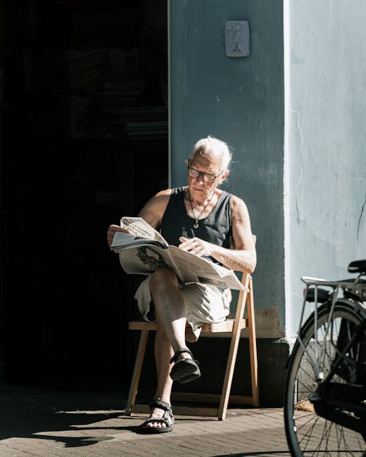 An elderly woman with short gray hair and glasses is seated on a small wooden chair outside a building with light gray wall surfaces. She is reading a newspaper, wearing a sleeveless black top, beige 